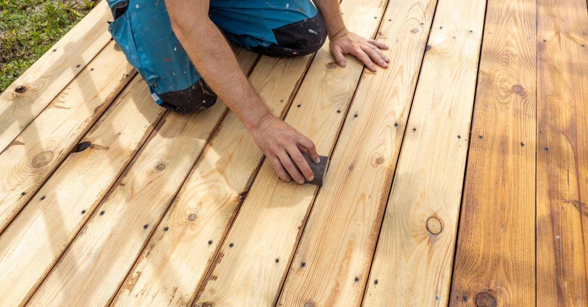 A contractor kneeling on an unfinished deck. The contractor is using a sanding block on the wooden planks of the deck.