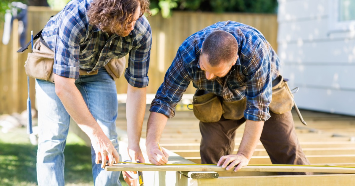 Two contractors working on a deck attached to a house. They are bent over the deck's frame placing a board of wood.