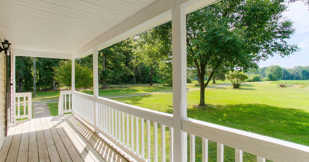 A wooden front porch with a white, wooden hand railing. There is a large green yard and a tree beyond it.