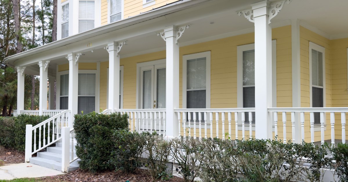 A yellow house's front porch with a white hand railing. The porch has stairs that lead down onto the home's yard.