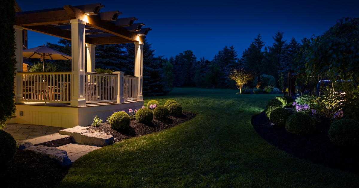 An outdoor pergola connected to a home, overlooking an expansive green lawn with luxurious landscaping at night.