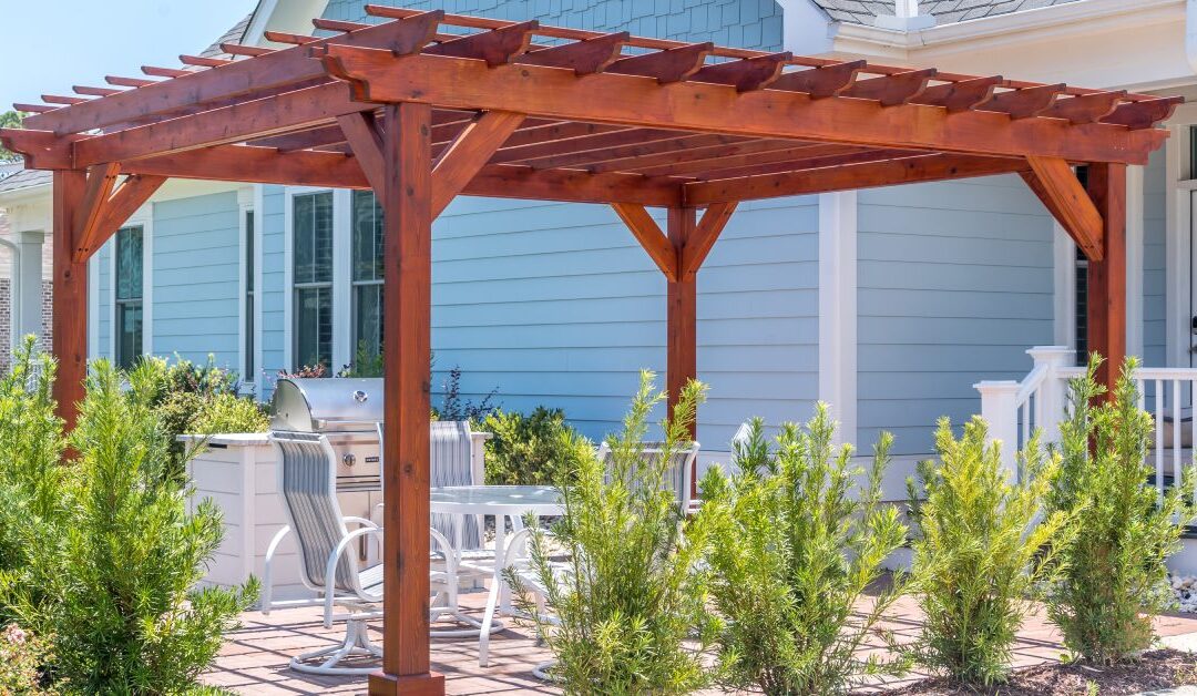 A wood pergola outside a home with blue siding. Bushes surround the structure and white patio furniture is underneath.