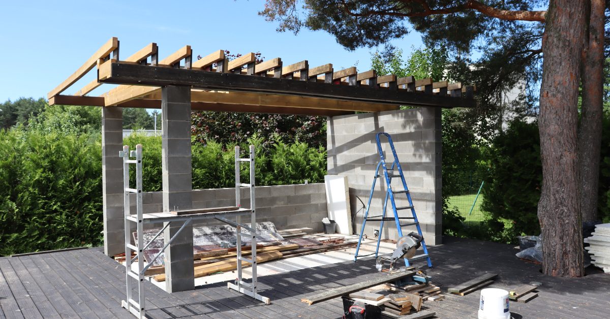 An outdoor pergola that's under construction has several ladders set up and scattered building materials.