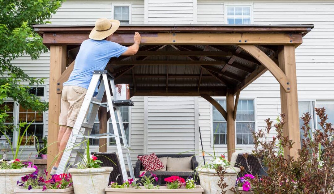 A man in a sun hat stands on a ladder to reach the roofline of an outdoor gazebo that is set up outside a home.