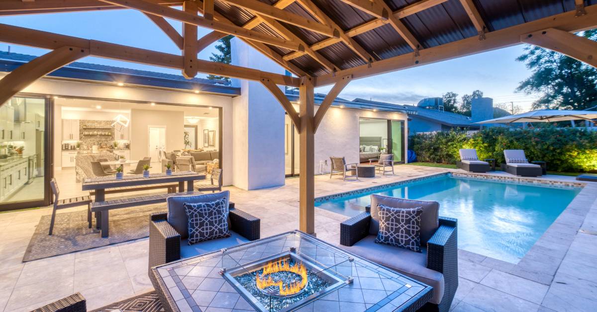 A fire table and chairs underneath a gazebo outside a large home. There is a swimming pool in the background.