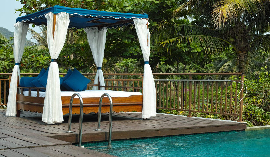 A wooden cabana with white drapes, blue throw pillows, and a blue cloth top, sitting on a wooden pool deck.