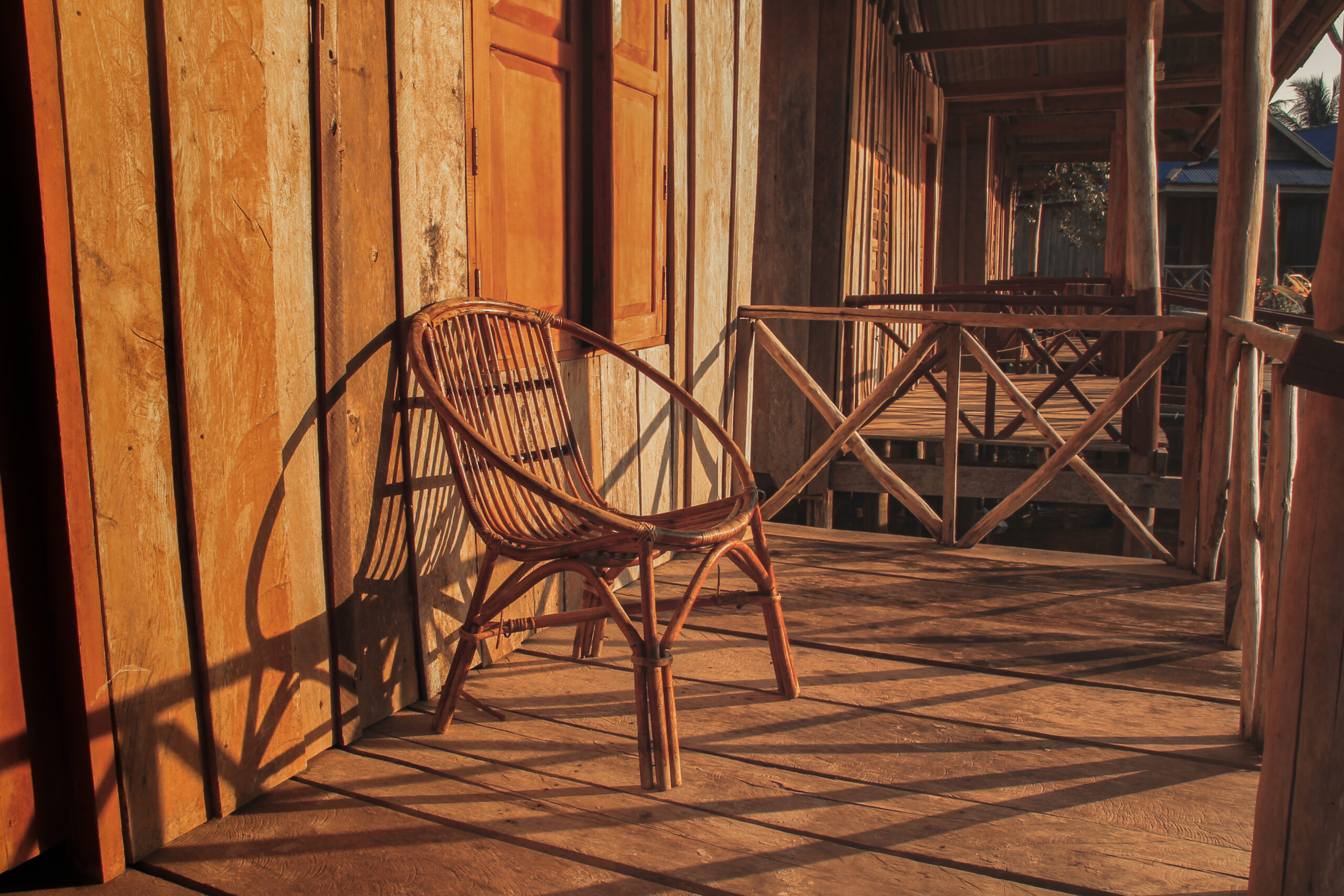 Sunlight stretches across a wooden porch with a round wood chair. A matching porch is in the background.